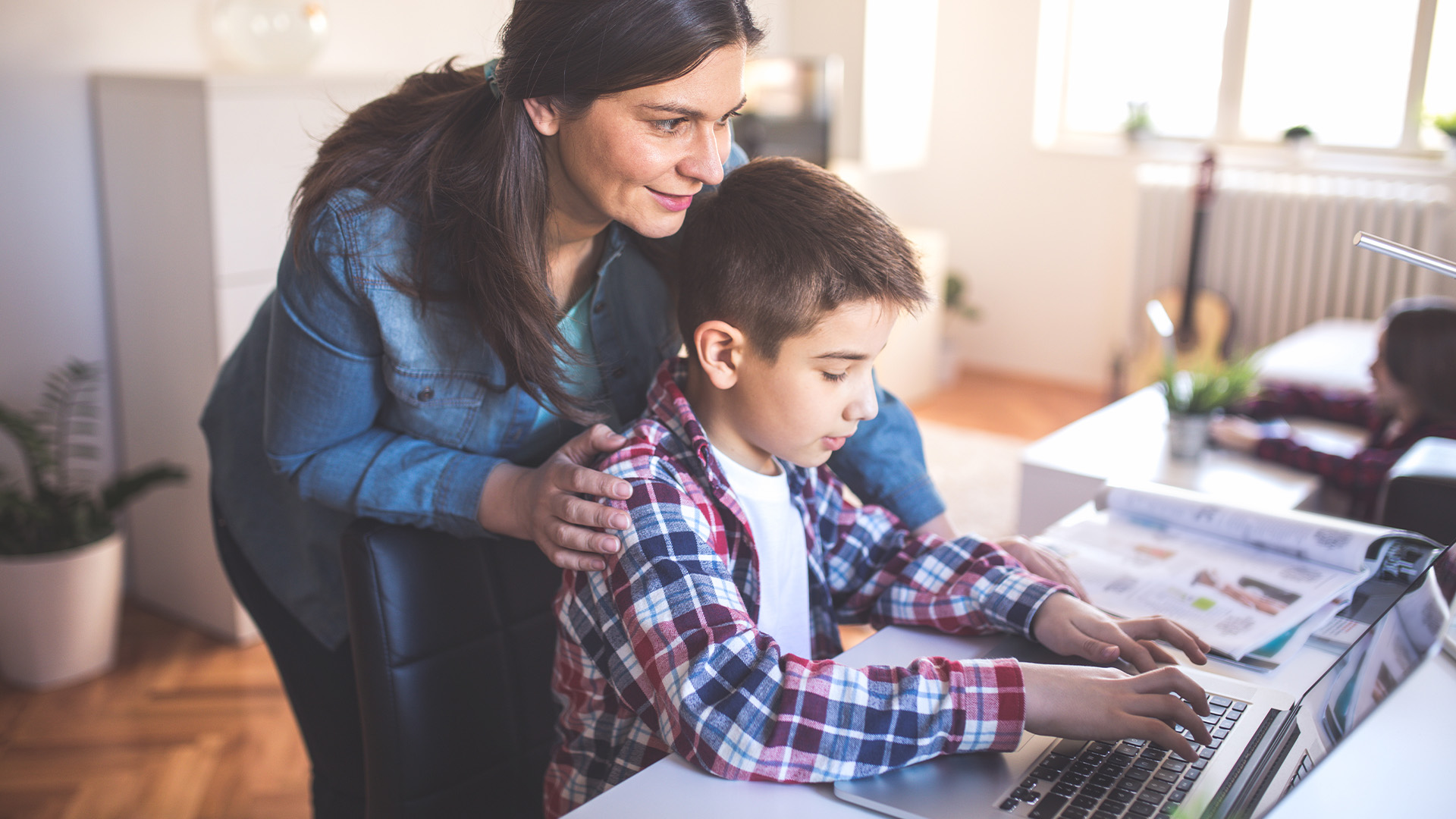 A mother leans over her son as he types on a laptop