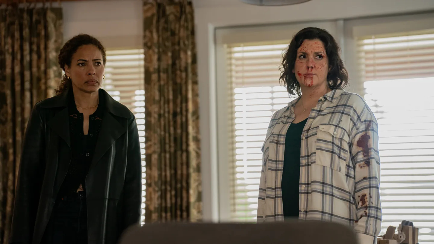 Tawny Cypress as Taissa, left, stands in a living room with Melanie Lynskey as Shauna, who has blood on her face and clothes, in 'Yellowjackets' season 3.