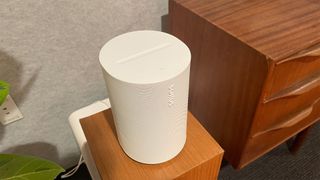 A white Sonos Era 100 speaker on a square wooden stand next to a wooden desk with drawers.