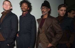 Four male models wearing looks from Oliver Spencer's collection. One model is wearing a brown high neck jumper and blue striped suit. Another model is wearing a dark blue shirt and trousers with another shirt in dark red underneath. Next to him is a model wearing a black hat, maroon top, black and white patterned trousers, brown jacket and brown coat on top. And the fourth model is wearing a brown high neck jumper, multicoloured striped trousers and a blue and brown jacket