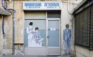 Barber shop front, grey concrete floor, light stone brick walls, black blind shutters to the right, grey drain pipe, faceless people cut-outs, smartly dressed male, barber cutting a smartly dressed customer placed in the window, side table cut-out to the left of the doorway