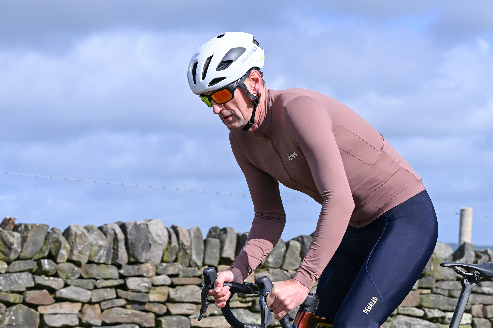 Man riding a bike wearing a white helmet, sunglasses and a long sleeved purple jersey in front of a drystone wall