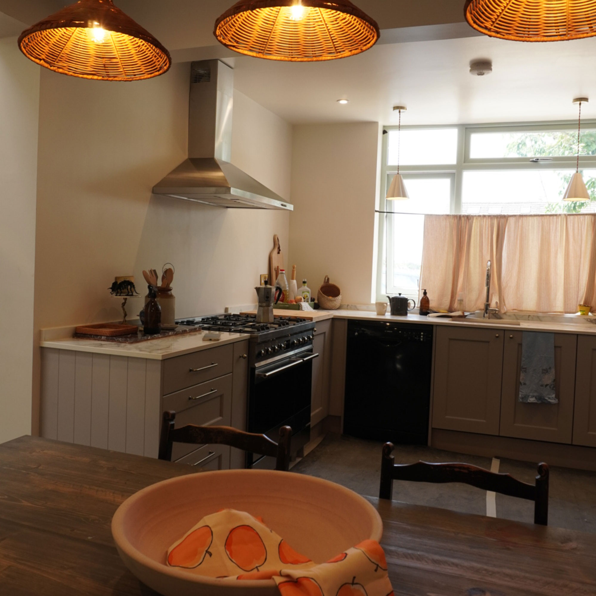 Kitchen neutral cabinetry, pale cafe curtain at window and with three wicker pendant lights above wooden table