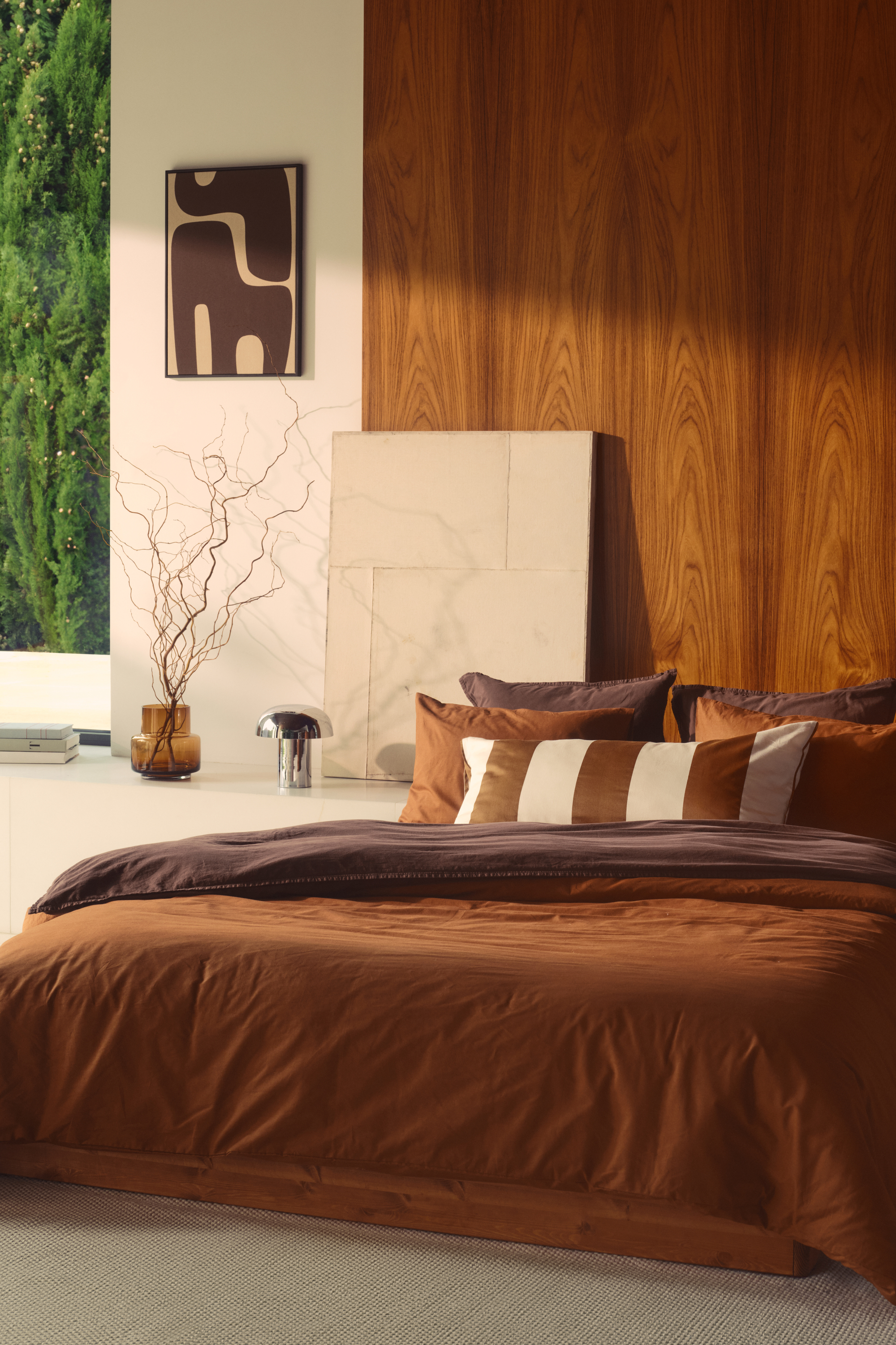 A modern bedroom with white and wooden walls. There is a bed that is low to the ground and has a burnt orange duvet and purple sheets. There is an orange and white striped throw pillow on the bed and a chrome mushroom lamp to the left.