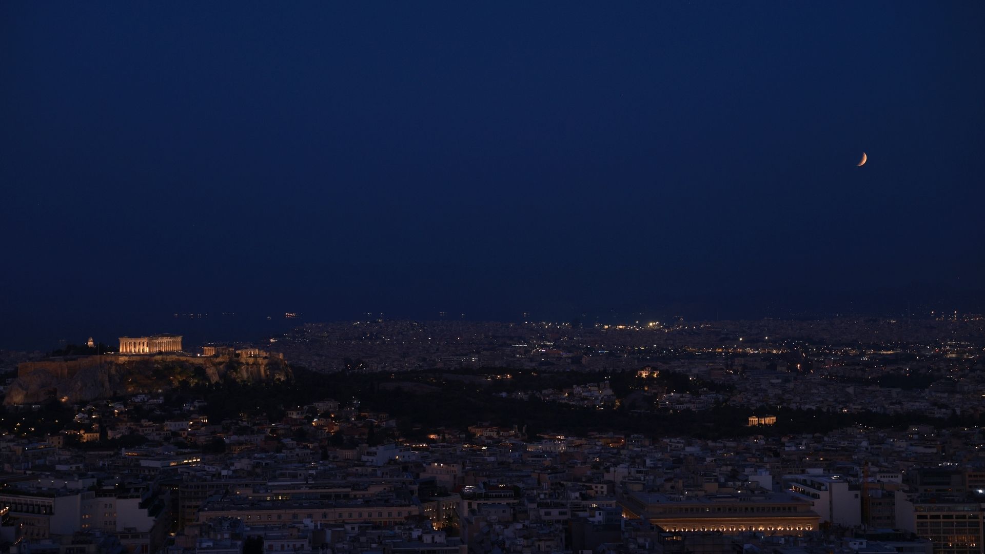 A photo of Athens at night. The eclipsed crescent of the moon is visible above the horizon, while the Parthenon is illuminated to the left.