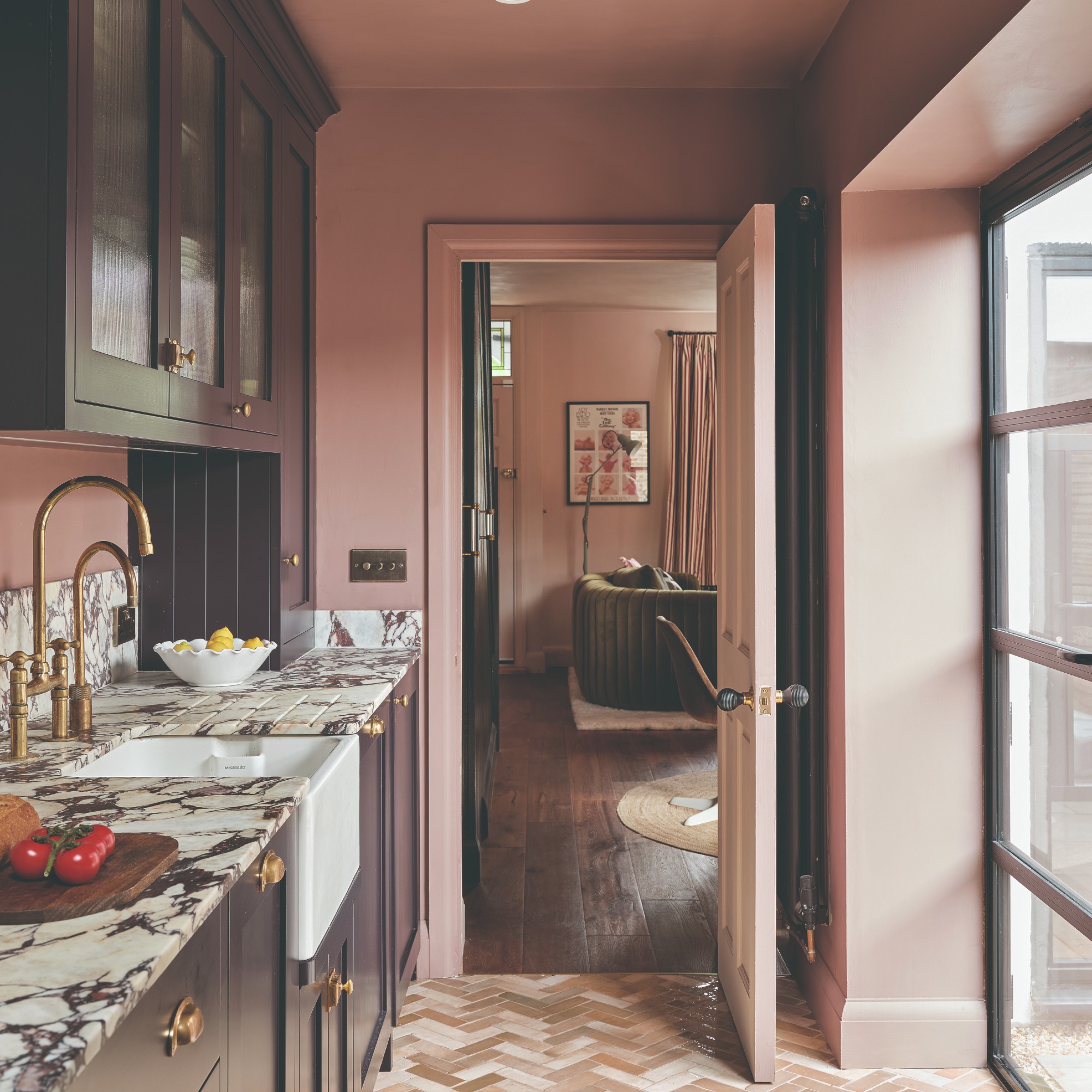 A pink kitchen painted in Farrow &amp;amp; Ball's Sulking Room Pink with dark purple cabinets and a view into a pink living room