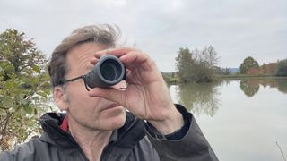 A man holding up the Vortex Solo R/T 8x36 monocular to his eye in front of a lake.