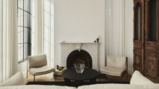 A small living room with sheer, white, recessed curtains hanging on the windows and walls. There is a marble fireplace and white furniture in the room.