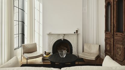 A small living room with sheer, white, recessed curtains hanging on the windows and walls. There is a marble fireplace and white furniture in the room.