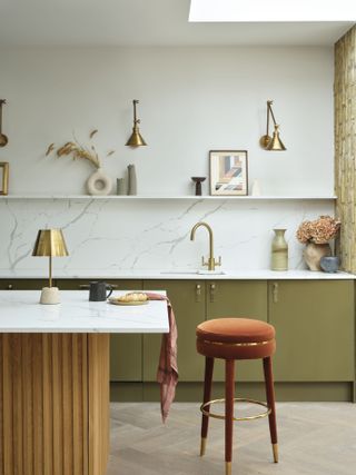 A kitchen with green cabinetry and brass wall sconces on the walls, plus a portable brass table lamp on the island