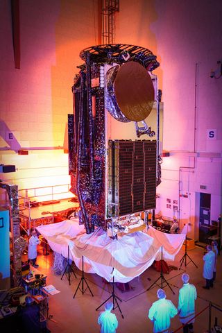 A towering satellite stands in a clean room facility, around hued lighting of oranges and pinks.