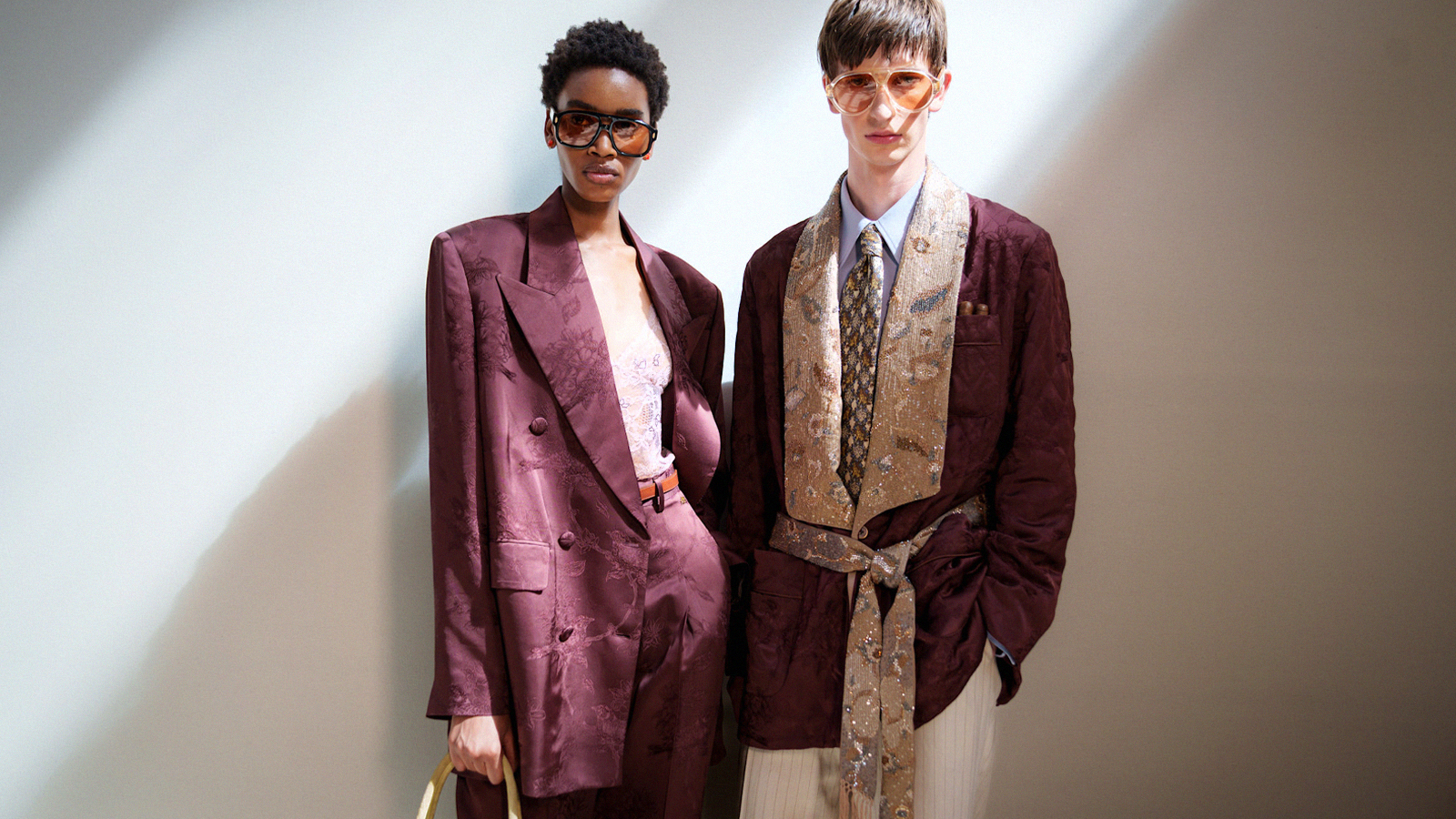 A female and male model stand in a beam of sunlight, the couple are both sharply dressed in relaxed tailoring, their suits are in a matching maroon fabric.