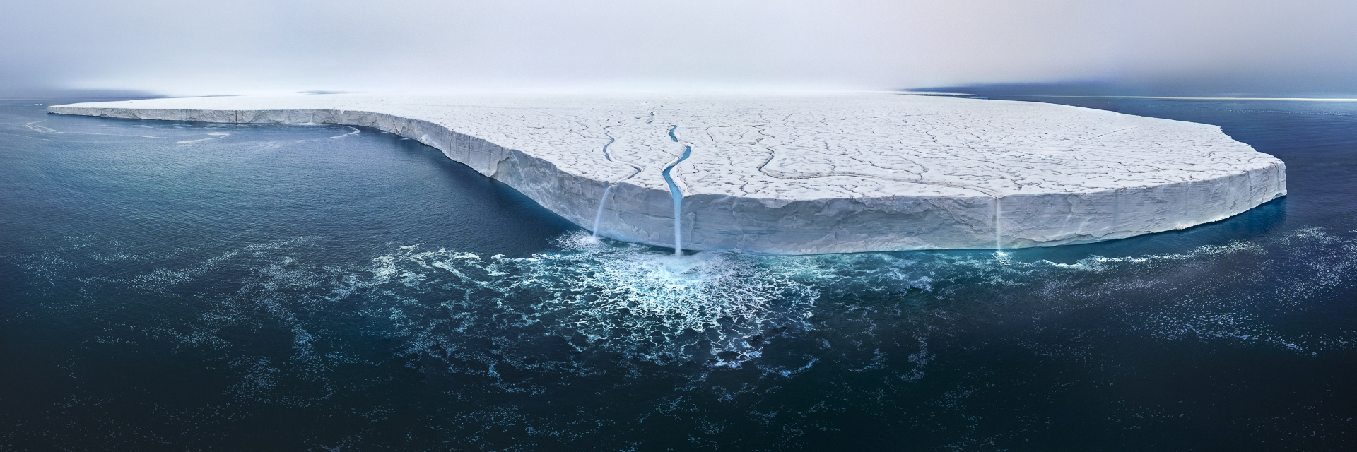 Aerial view of a massive iceberg with a smooth, white surface and prominent cracks, surrounded by deep blue ocean waters