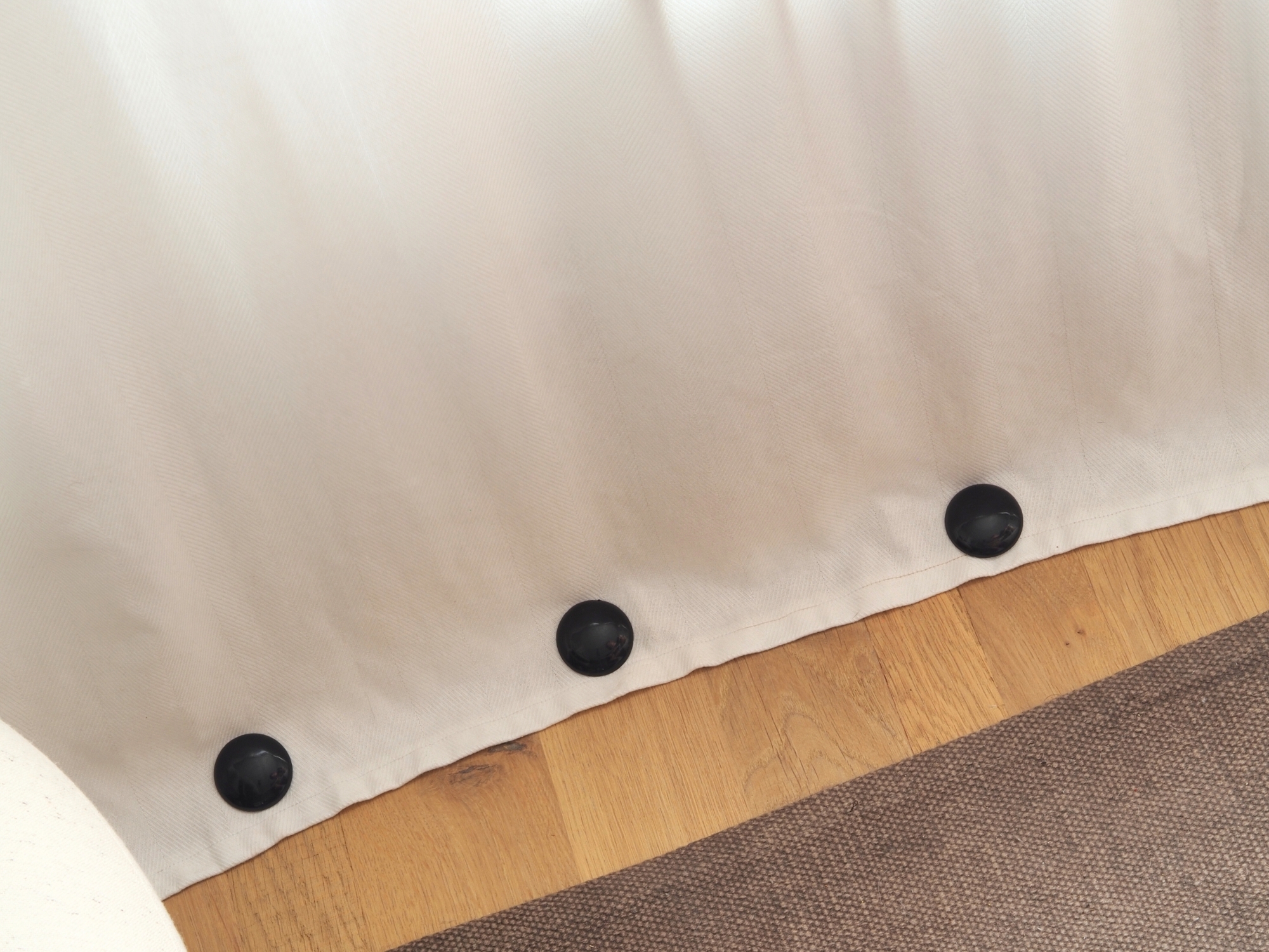 Close-up image of three black curtain magnets on the bottom edge of a white curtain. The hardwood floor is in view, and there is a gray/mocha rug.
