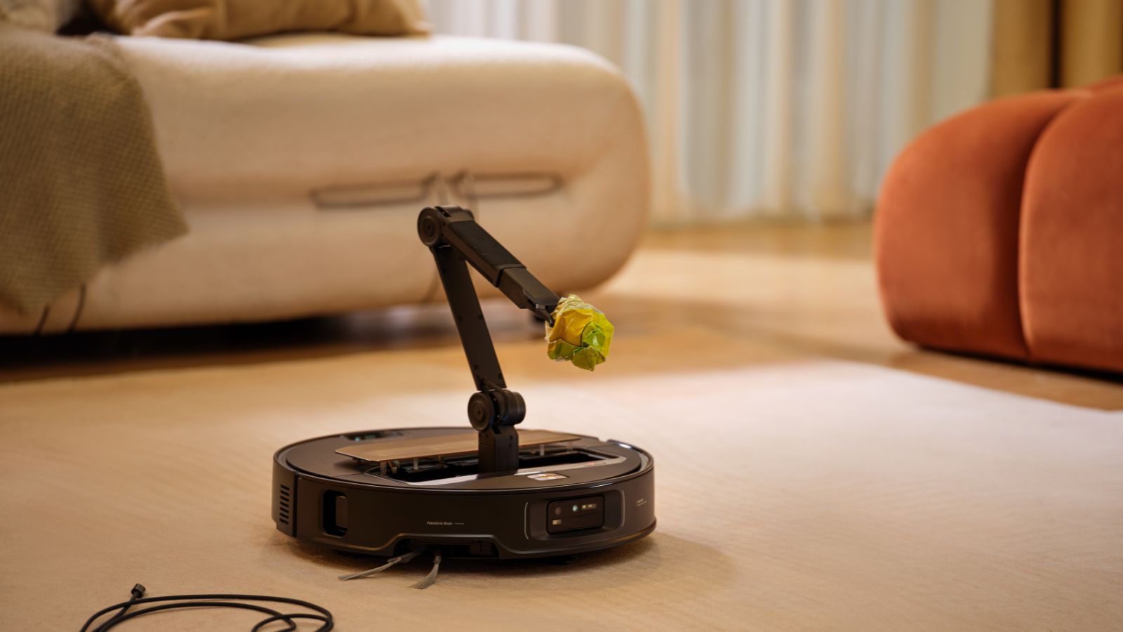 Roborock Saros Z70 on rug in front of orange velvet sofa in warm-toned living room, holding a crumpled ball of paper using its mechanical arm.