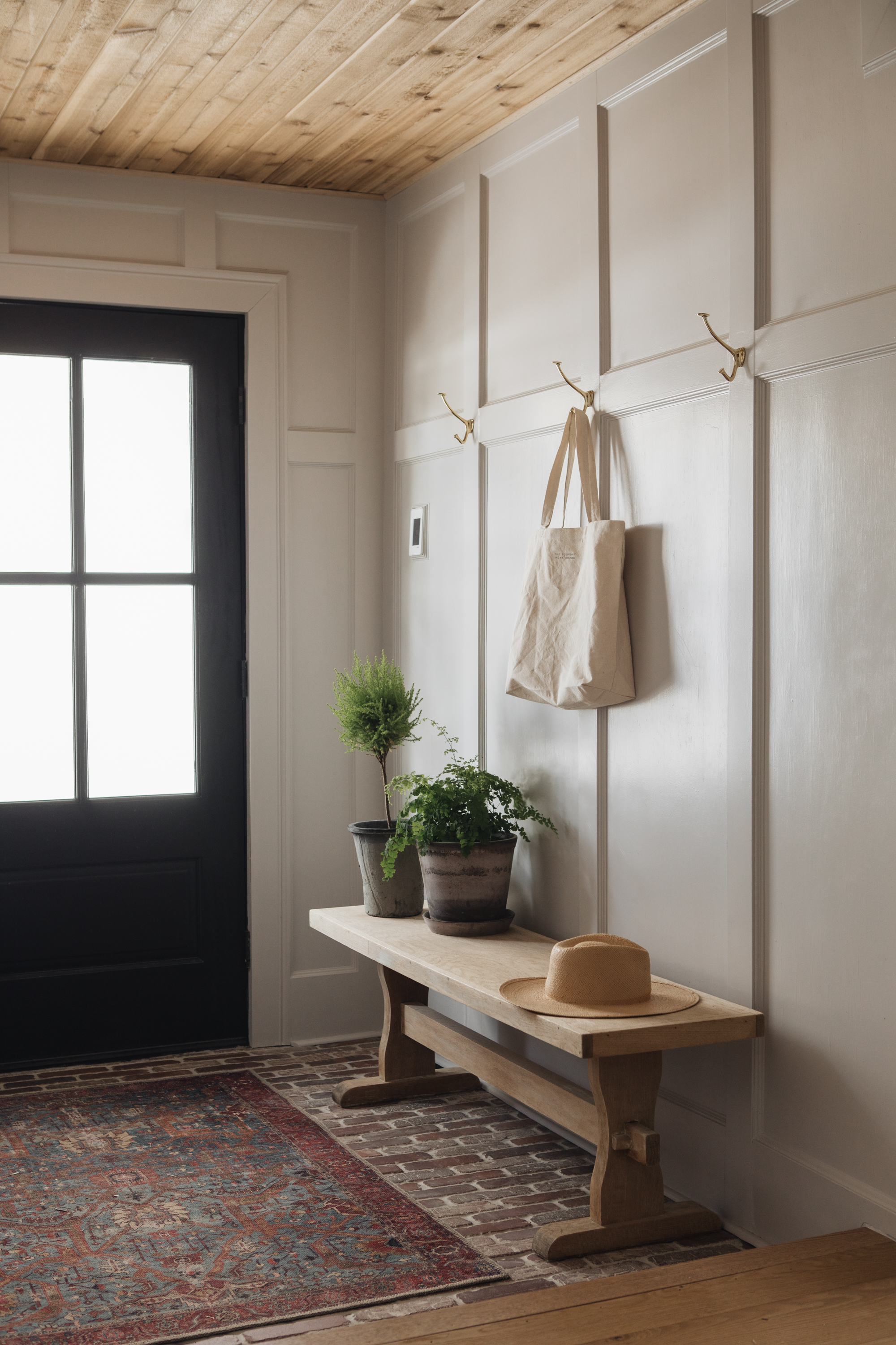 Image of a bright entry way painted in an off-white/green color. There is a wooden bench with plant pots on it and three hooks hanging on the wall above.