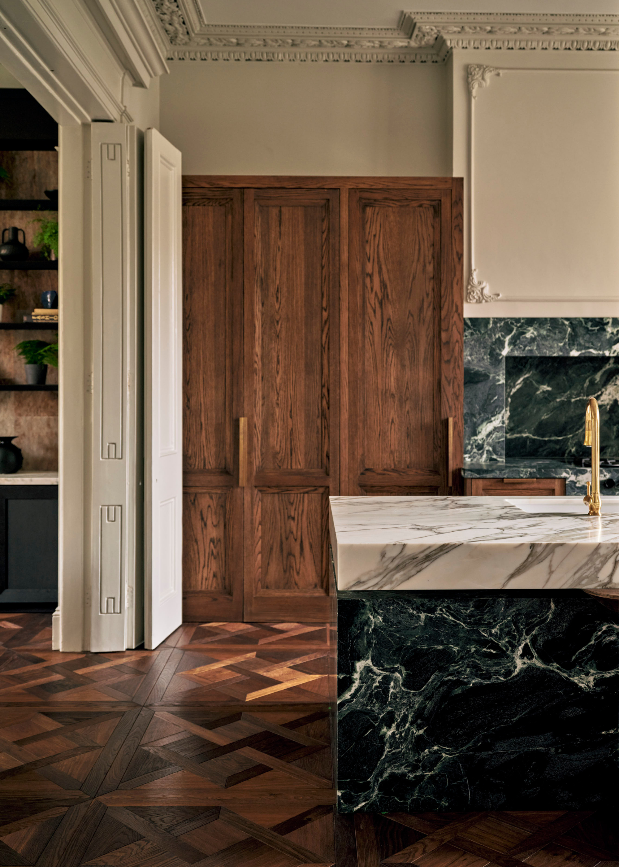 A kitchen with wood flooring and cabinets beside a green stone island with a white stone top and gold hardware