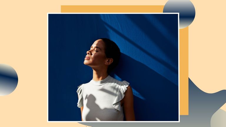 woman standing against blue wall with the sun shining on her face on an orange background