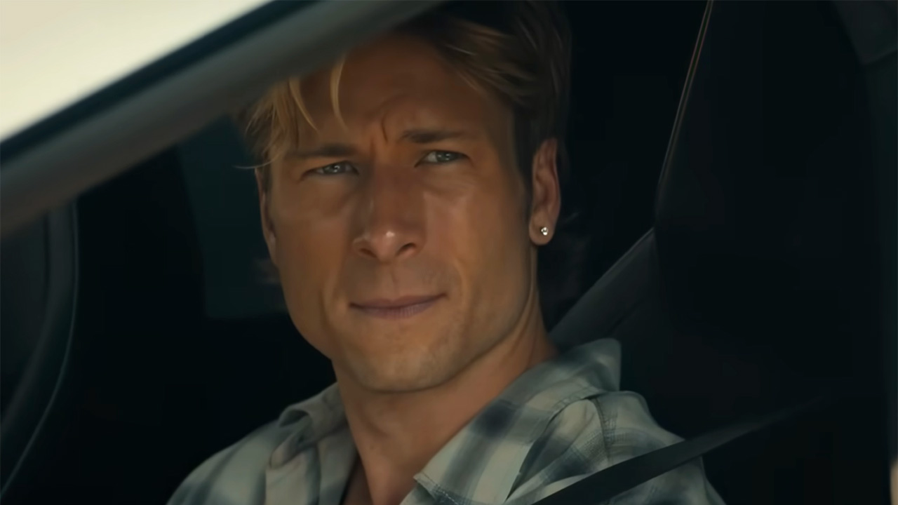 Glen Powell with a diamond stud and plaid shirt on sitting in a car and staring out a window in Chad Powers.
