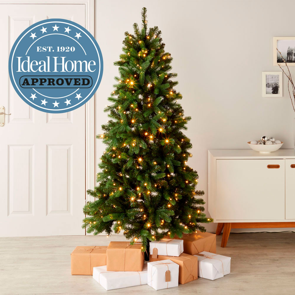 A green Christmas tree decorated with lights and gifts underneath in a white living room. Ideal Home approved logo overlaid at the top.