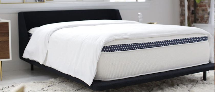 The image shows a white mattress with a navy blue trim placed on a dark blue bed foundation in a white bedroom