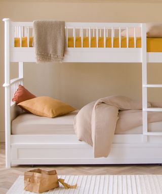 A white wooden bunk bed, one with a mustard yellow mattress and one with a beige mattress with yellow and red throw pillows on top of it, with a white rectangular rug on the floor next to it with a woven storage box