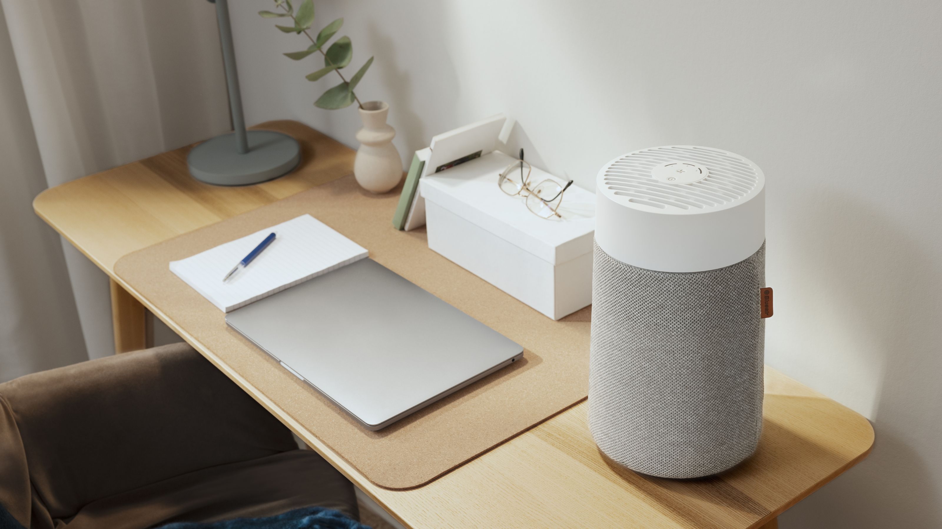 Blueair air purifier sitting on wooden desk, next to a laptop and other desk items