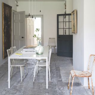 small narrow dining room with stone floor, white table and chairs artwork, bare bulb pendants, grey painted door, metal antique chair
