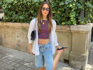 Dutch fashion influencer Luna Isabella on the street during summer wearing an oversize white shirt open over a cropped striped tank, long denim shorts, kitten-heeled black flip-flops, rectangular sunglasses, a Chanel shoulder bag, and a gold heart necklace.