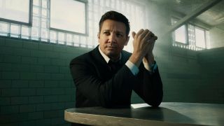 Jeremy Renner sitting at a table with his hands clapsted together while his elbows rest on the table.