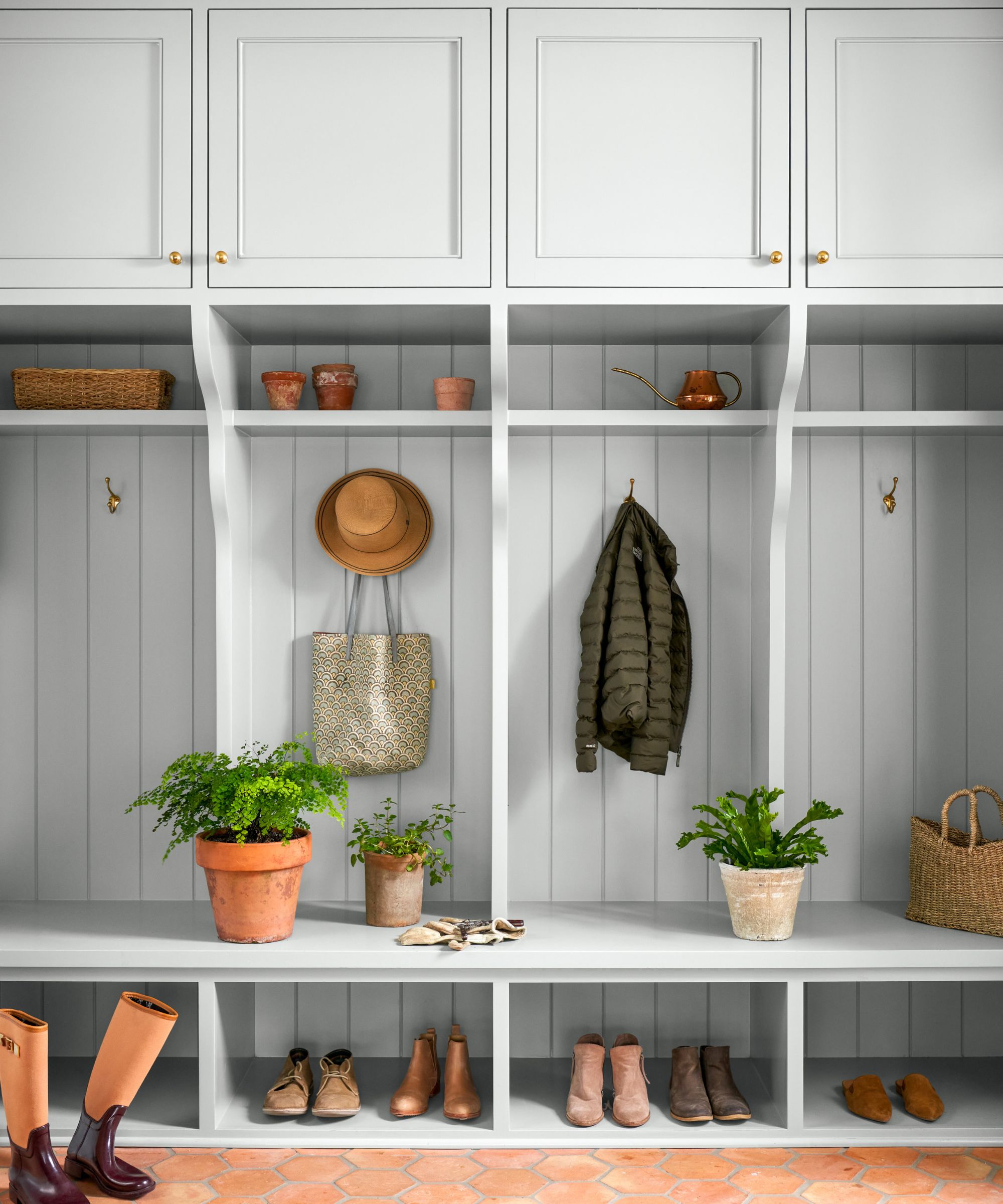 A mudroom with light gray joinery, including open shelving, a seating bench, and closed cupboards. Various shoes, bags, and coats are stored, with terracotta flooring.