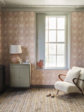 bedroom with pink wallpaper, grey woodwork, upholstered armchair, small chest, lamp, stripe carpet
