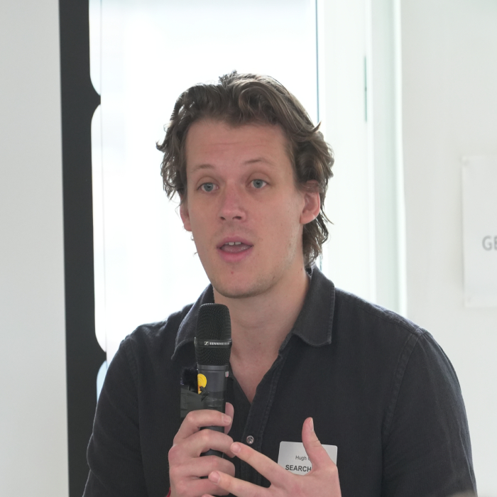 male with medium length brown hair wearing black polo shirt talking with microphone in hand