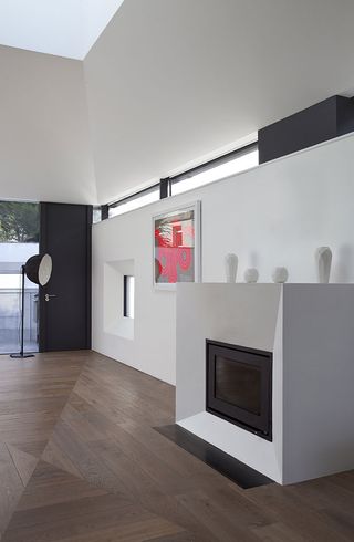 A white wall with an fire place. White framed painging on the wall. Dark wooden flooring and white ceiling. Black standing lamp