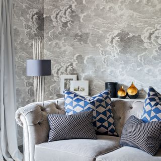 grey living room with a grey patterned wallpaper and grey sofa with blue geometric patter cusions and a modern silver and blue floor lamp