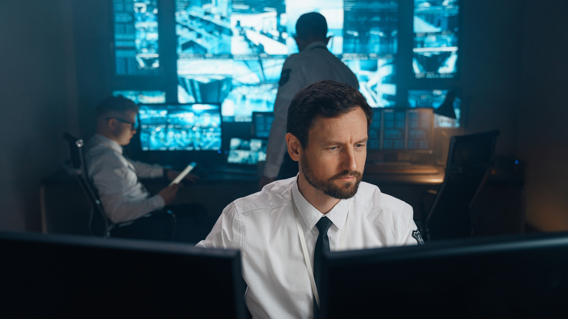 A close-up shot of a military or cybersecurity worker in a shirt and tie, sat in front of two monitors, with two glowing blue screens showing security camera footage on them in the background, partially obscured by two other workers.