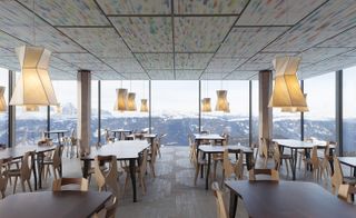 Alternative view of AlpiNN restaurant featuring multiple tables, chairs, low hanging asymmetric pendant lights and wooden flooring. The ceiling is multicoloured and views of the mountains can be seen through the floor to ceiling windows