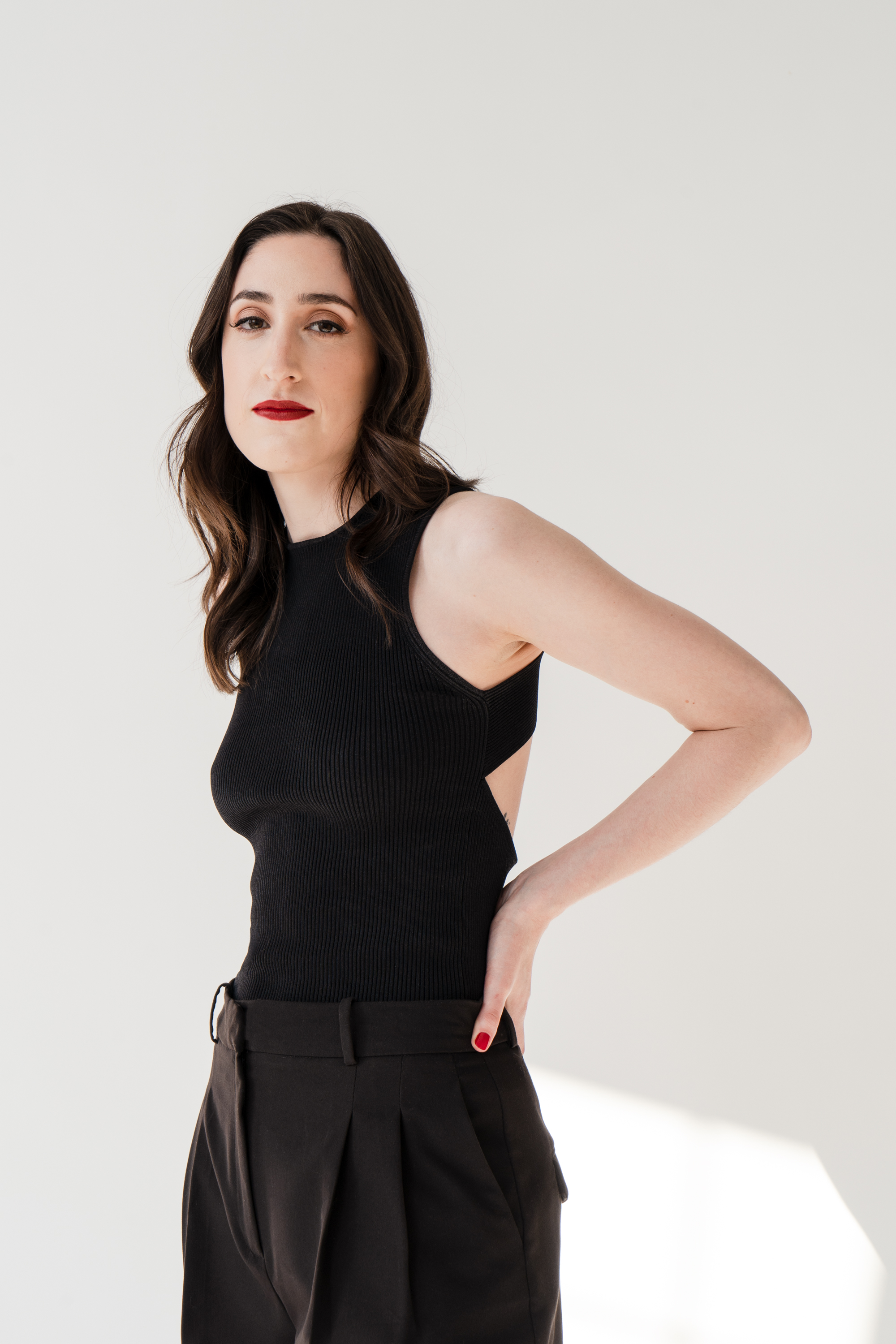 A portrait of a woman (Izzy Custodio) in a black sleeveless top and slacks, standing in front of a white backdrop.