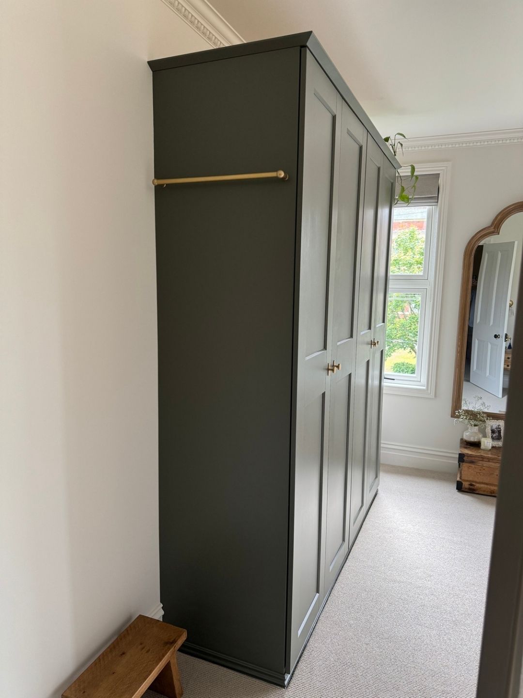Side view of a sage green wardrobe in a white room that has white carpeting and a window and mirror on the far wall.