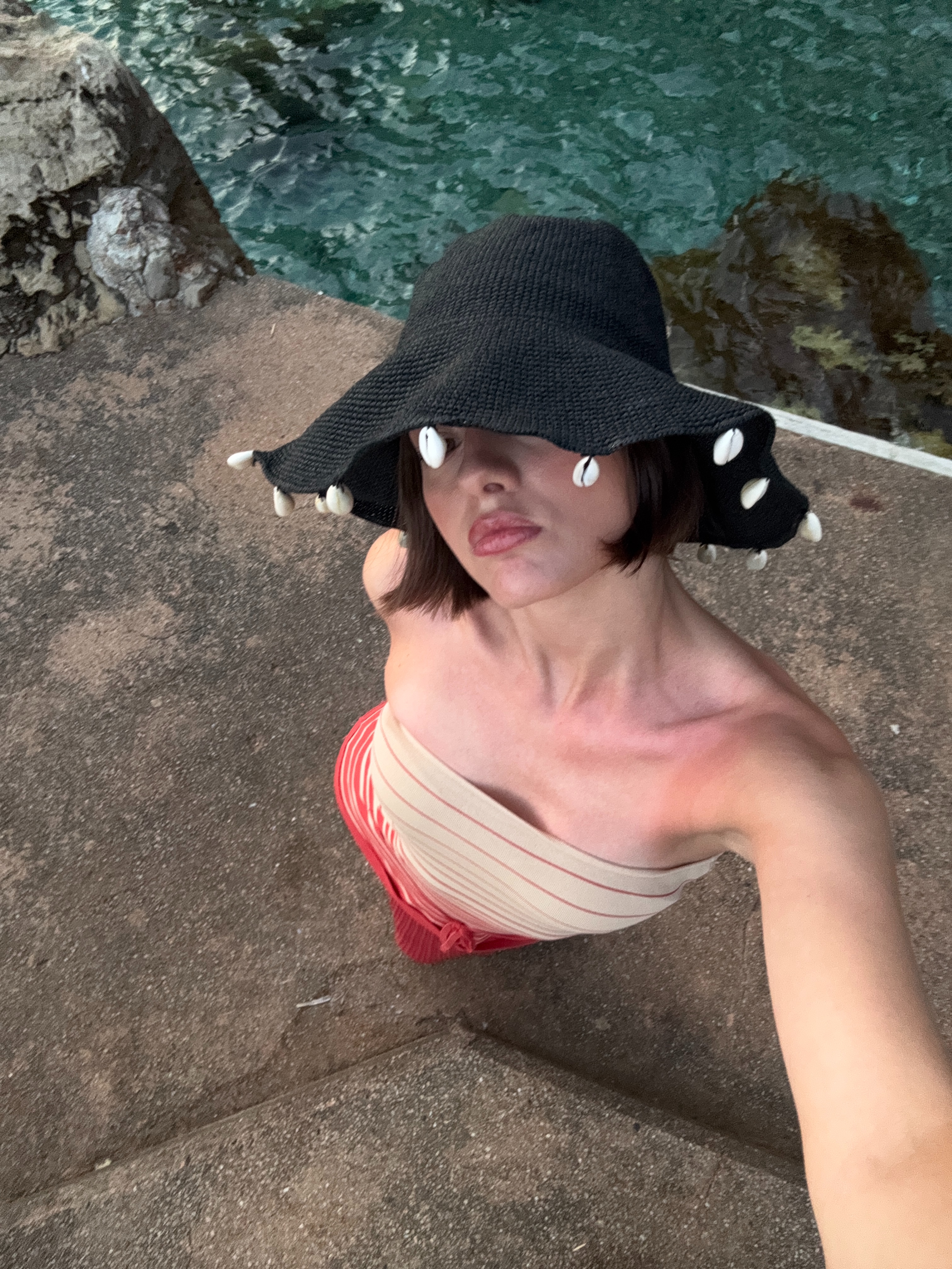 A woman standing by a rocky coastline wearing a shell-adorned hat.