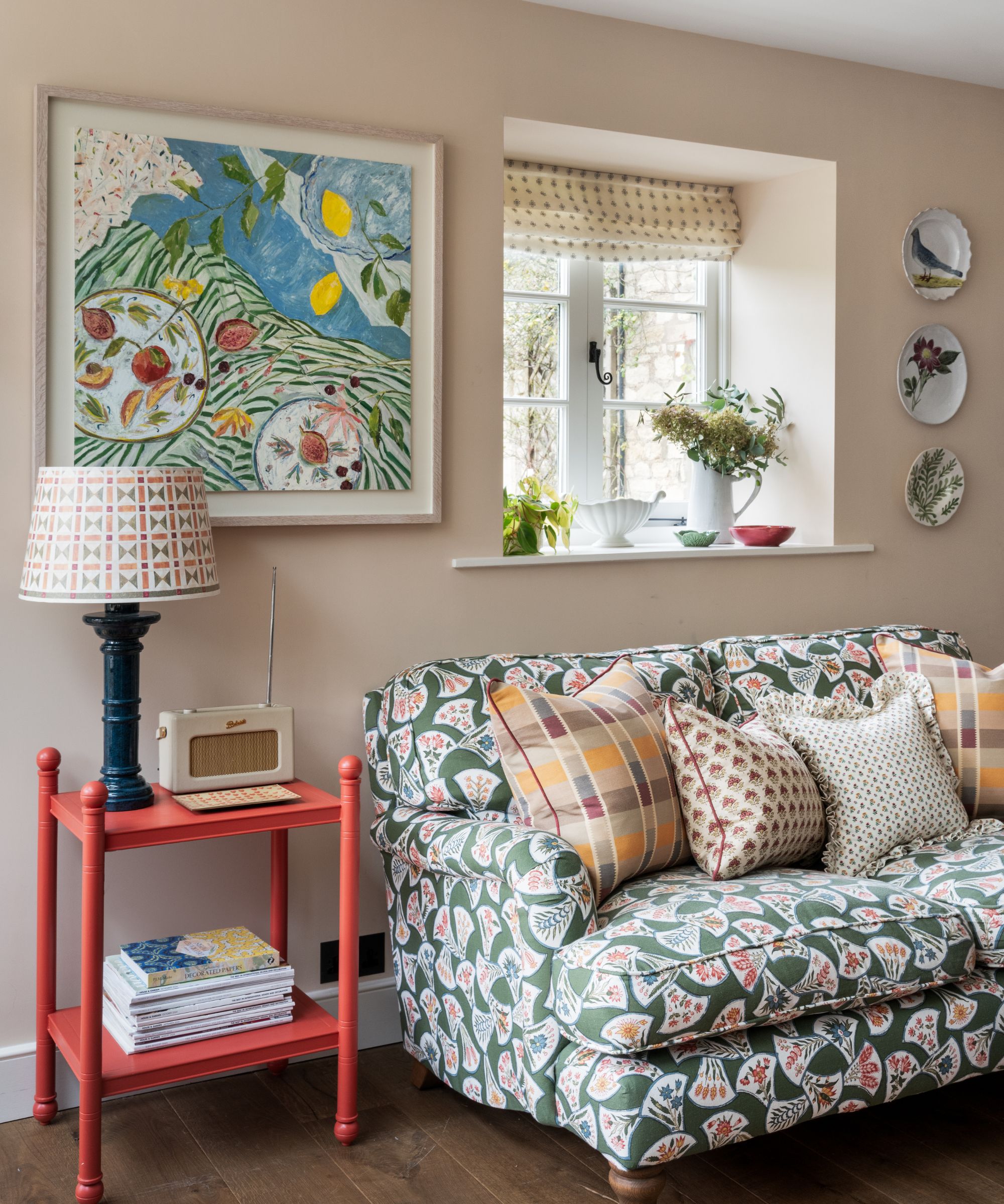Living room with pale pink walls, plates and art on the wall, green floral sofa with an array of colourful cushions and a red-painted side table with a lamp and radio