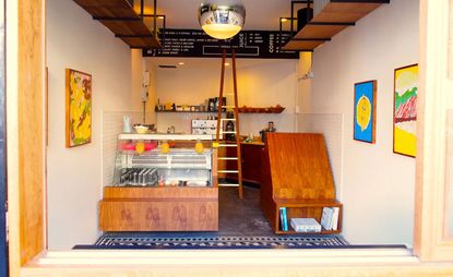 Interior view of Max Bagels featuring light coloured walls, a chrome sphere light, high level wooden shelving, colourful art on the walls, wooden counters, a black price board with white writing and view of the kitchen area