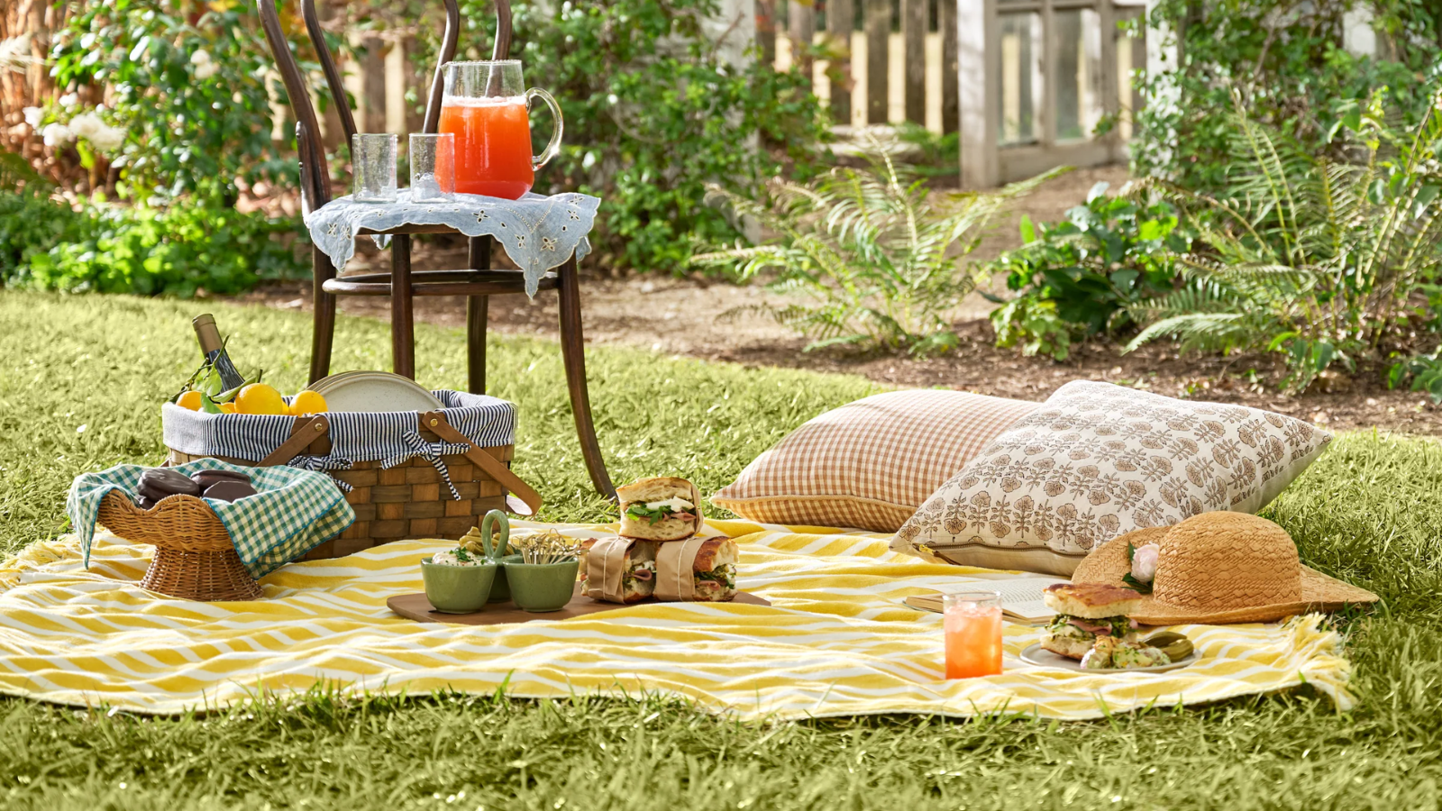 a picnic scene with a yellow blanket, pillows, snacks, and a chair with a jug and glasses on it