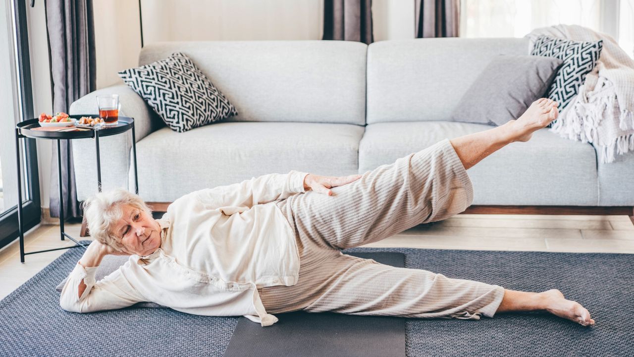 woman dressed in cream loose top and trousers lying on her side facing the camera with her top leg raised. she's on a grey carpet with a light grey sofa behind her. 