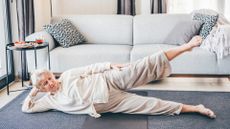 woman dressed in cream loose top and trousers lying on her side facing the camera with her top leg raised. she's on a grey carpet with a light grey sofa behind her. 