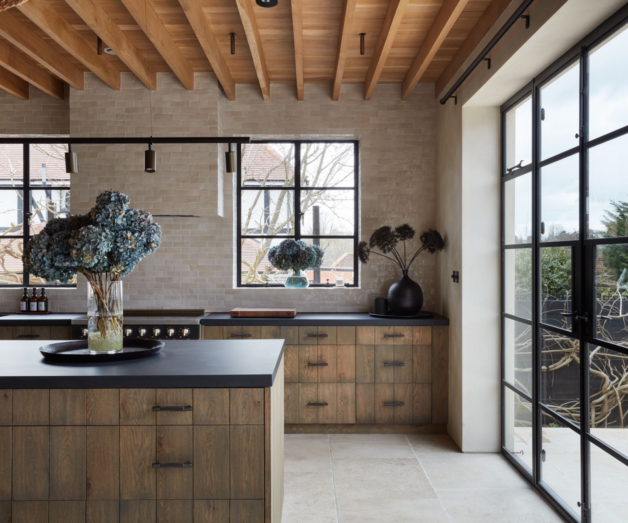 Light-filled kitchen with timber cabinets, stone walls and exposed beams. A large island faces black-framed windows overlooking a garden.