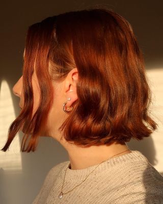 Woman with short red bob haircuit side profile facing camera, wearing diamond earrings and simple gold hoop earrings.