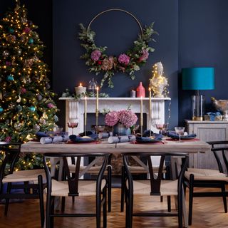 blue purple and pink theme living room with dark blue walls white mantle wooden and black dining table and a decorated christmas tree with matching wreath hanging on the wall