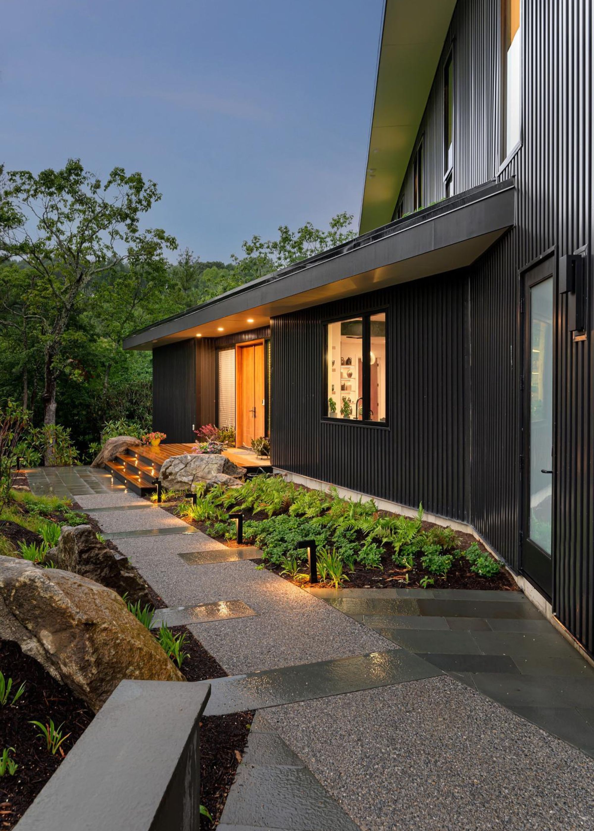 A garden pathway light up at night with path lights along the side of a house clad in black paneling with spotlights above the wooden front door and greenery all around