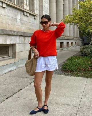 Female fashion influencer Sasha Mei poses wearing oversize sunglasses, a bright red sweater, tan suede bag, white cotton shorts, and black Mary Jane flats